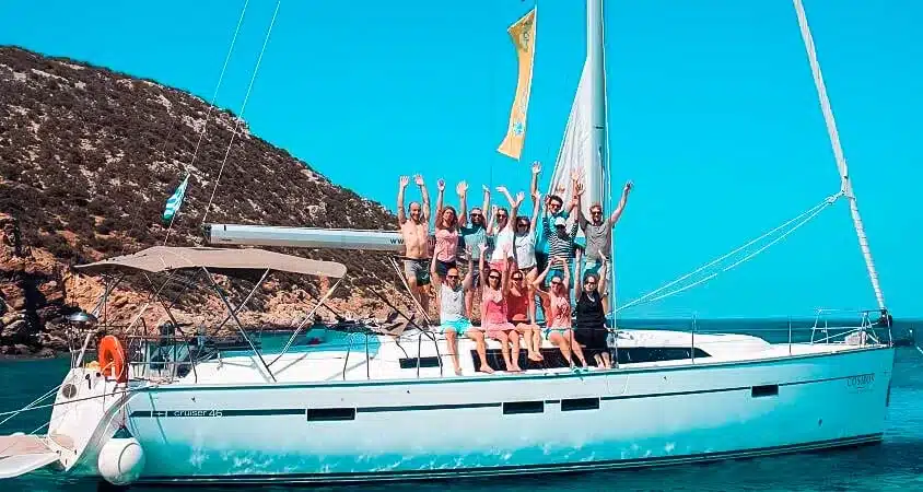 Eine Gruppe glücklicher Menschen hebt ihre Hände auf einer Segelyacht, die in einer ruhigen blauen Bucht mit felsigen Hügeln im Hintergrund vor Anker liegt.