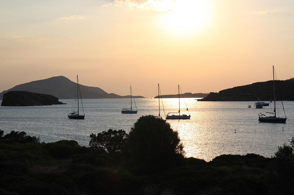 Sonnenuntergang über einer ruhigen Bucht mit Silhouetten verankerter Segelyachten und sanften Hügeln im Hintergrund. Das Sonnenlicht spiegelt sich im Wasser und schafft eine ruhige Szene.