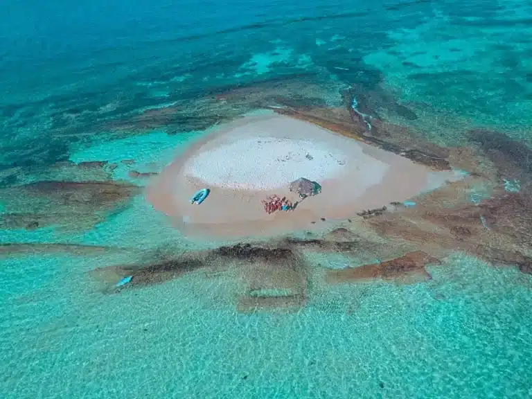 Luftaufnahme einer kleinen, herzförmigen Sandinsel, umgeben von klarem, türkisfarbenem Wasser. Ein paar Menschen und Boote sind zu sehen, perfekt für einen ruhigen Segelurlaub.