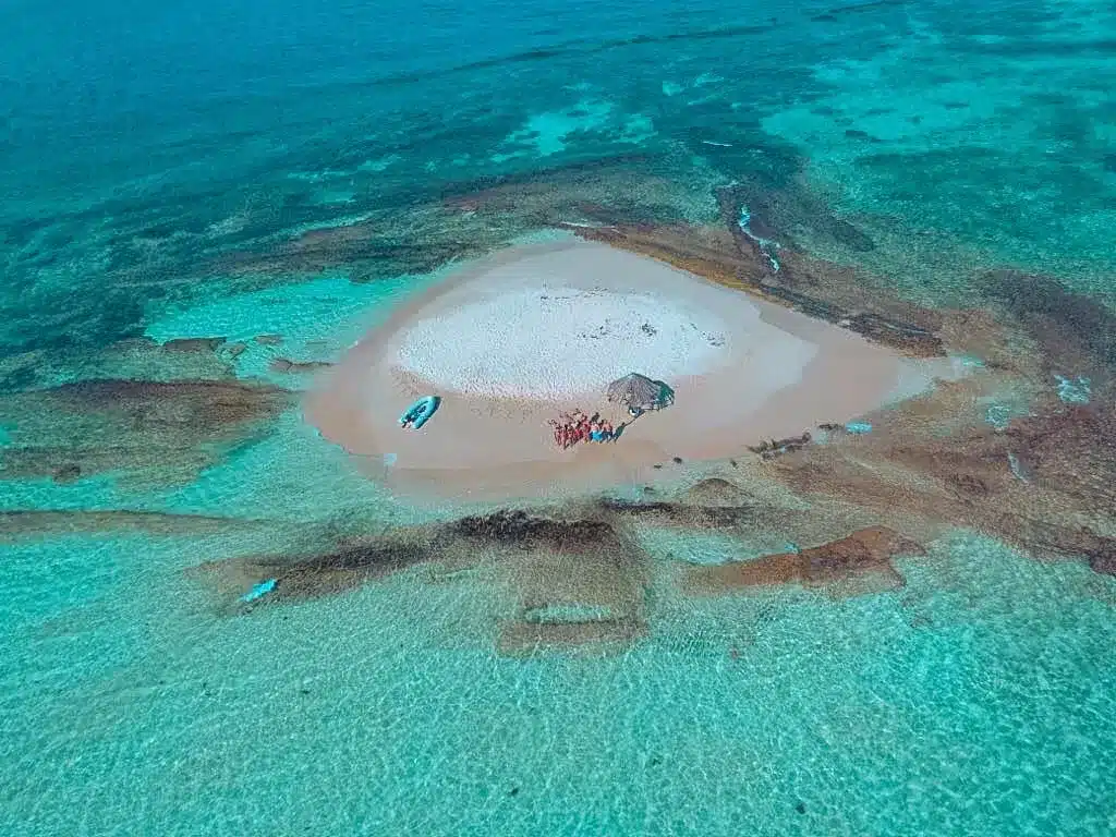 Luftaufnahme einer kleinen Sandinsel, umgeben von kristallklarem, seichtem Wasser. Zu sehen sind einige Boote, darunter eine Segelyacht, und Menschen, die die leuchtenden Meeresfarben hervorheben.