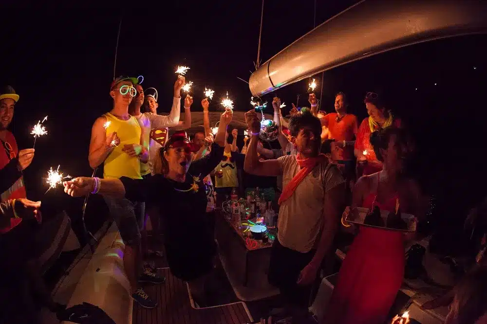 Eine Gruppe von Menschen mit Party-Accessoires hält während einer nächtlichen Segelreise auf einem Boot Wunderkerzen in der Hand und sorgt so für eine festliche und fröhliche Atmosphäre.