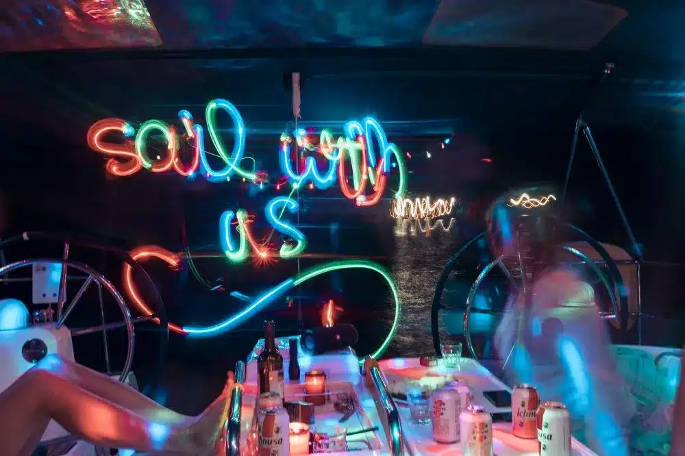 Leuchtende Neonlichter zeigen den Schriftzug „Soul with us“ in einem dunklen Raum mit spektralem Unschärfeeffekt. Alkoholische Getränke sind auf einem Tisch im Vordergrund der Segelyachtkabine verstreut.