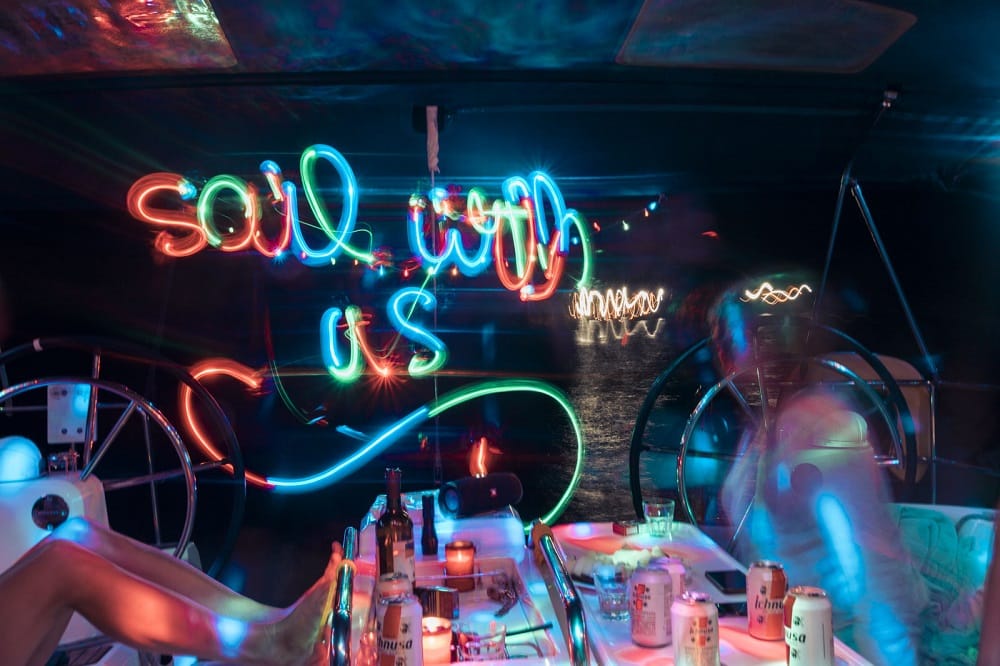 Eine lebhafte Partyszene auf einem Boot mit Neonlichtern, in denen „Segel mit uns“ steht, über einer Gruppe von Menschen, die ihren Segeltörn genießen, mit Getränken und festlicher Beleuchtung, was eine lebhafte Nachtleben-Atmosphäre schafft