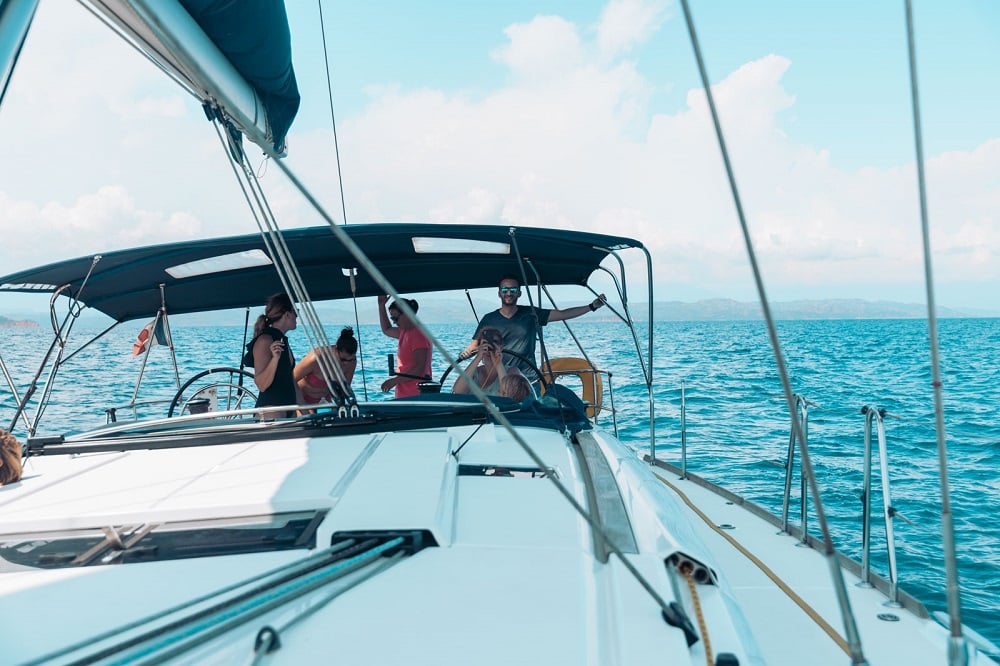 Eine Gruppe von Menschen genießt einen sonnigen Tag auf einem Segelboot und segelt auf klarem, blauem Wasser mit sichtbarer Küstenlinie in der Ferne. Der Blick geht vom Bug zum Heck.
