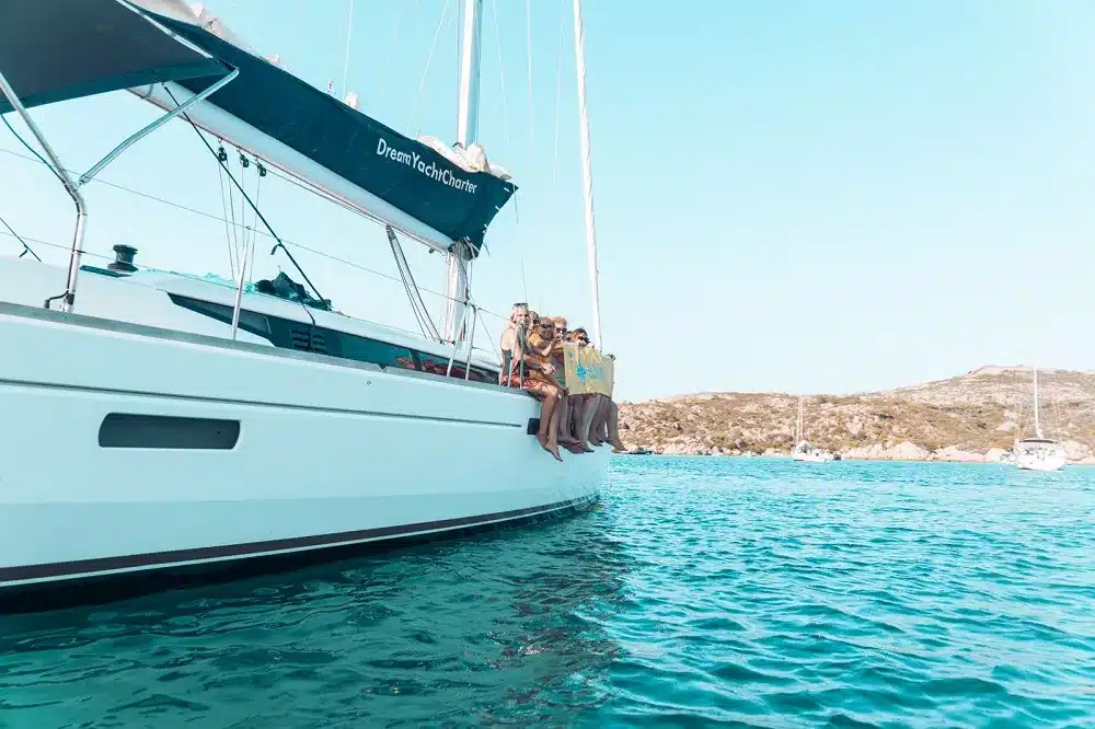 Eine Gruppe von Menschen genießt einen sonnigen Tag auf dem Deck einer Segelyacht, die im klaren, türkisfarbenen Wasser nahe einer felsigen Küste vor Anker liegt.