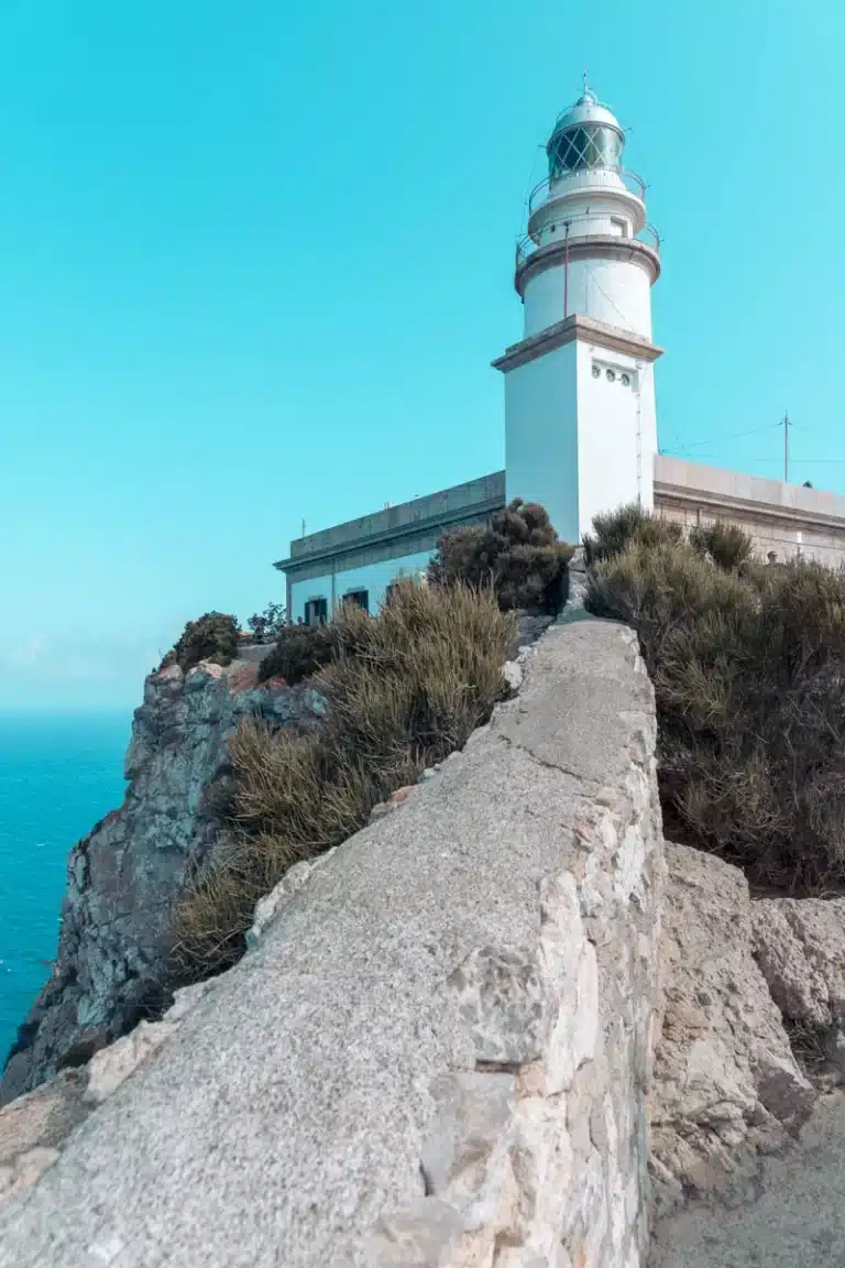 Ein hoher weißer Leuchtturm auf einer Klippe mit Blick auf das Meer, mit einem klaren blauen Himmel darüber und Flecken grüner Büsche auf dem felsigen Hang in der Nähe der Segelyacht.