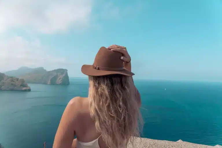 Eine Frau mit Hut blickt von einem hohen Aussichtspunkt an der Küste auf ein ruhiges, türkisfarbenes Meer, in der Ferne sind neblige Klippen zu sehen, während sie ihren nächsten Segeltörn plant.