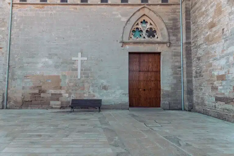 Eine Außenwand einer alten Steinkirche mit einer großen Holztür und einem kleinen weißen Kreuz, davor eine Bank unter einem dekorativen Bogenfenster.