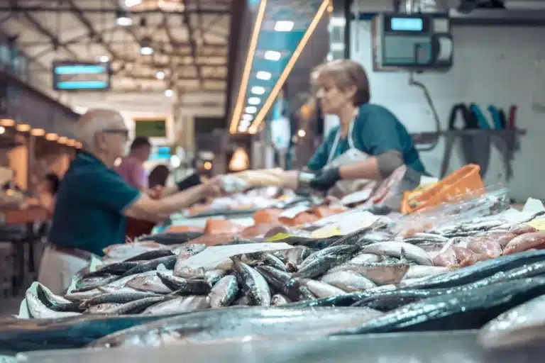 Eine ältere Frau kauft Fisch an einem Marktstand und gibt dem Verkäufer Geld. Im Vordergrund ist eine Segelyacht mit einer Auswahl an frischem Fisch gut sichtbar ausgestellt.