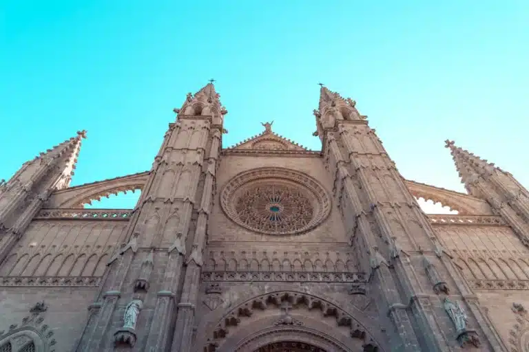 Die Fassade einer großen gotischen Kathedrale mit detaillierten architektonischen Gravuren, zwei hoch aufragenden Türmen und einem großen runden Buntglasfenster unter einem klaren blauen Himmel, perfekt zum Segeln.