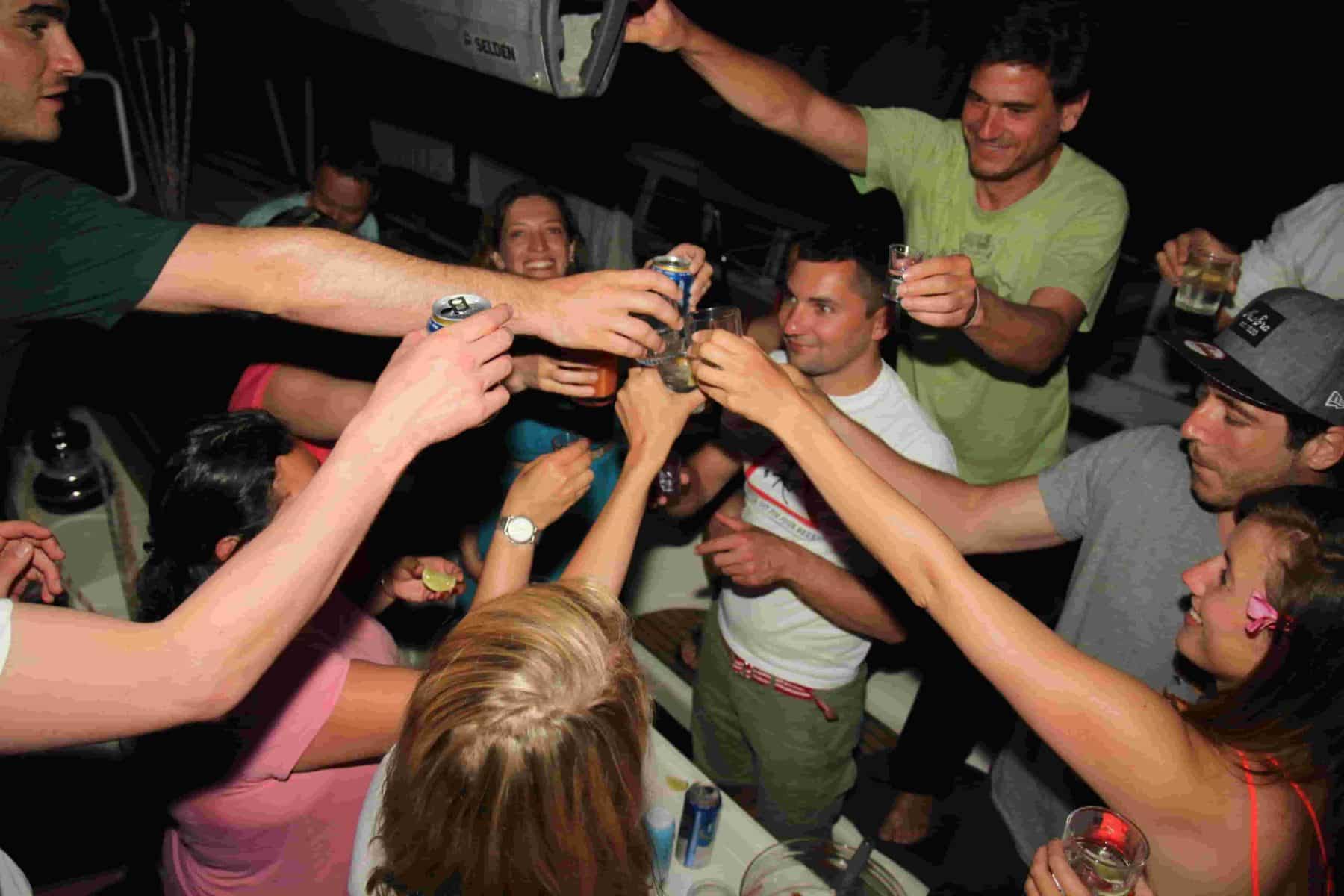 Eine Gruppe von Menschen stößt nachts fröhlich mit Getränken auf einer Segelyacht an.