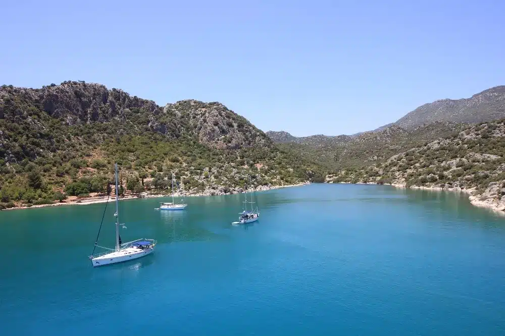 Eine ruhige Bucht mit tiefblauem Wasser, umgeben von üppigen grünen Hügeln, mit drei Segelbooten, die friedlich unter einem klaren Himmel vor Anker liegen, perfekt für einen Segeltörn.