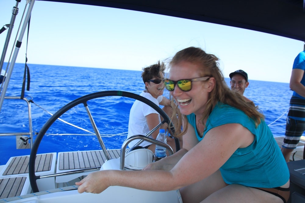Eine fröhliche junge Frau mit roten Haaren, Sonnenbrille und blaugrünem Oberteil steuert auf ihrer Segelreise ein Boot mit großem Steuerrad. Im Hintergrund sieht man Passagiere, die den