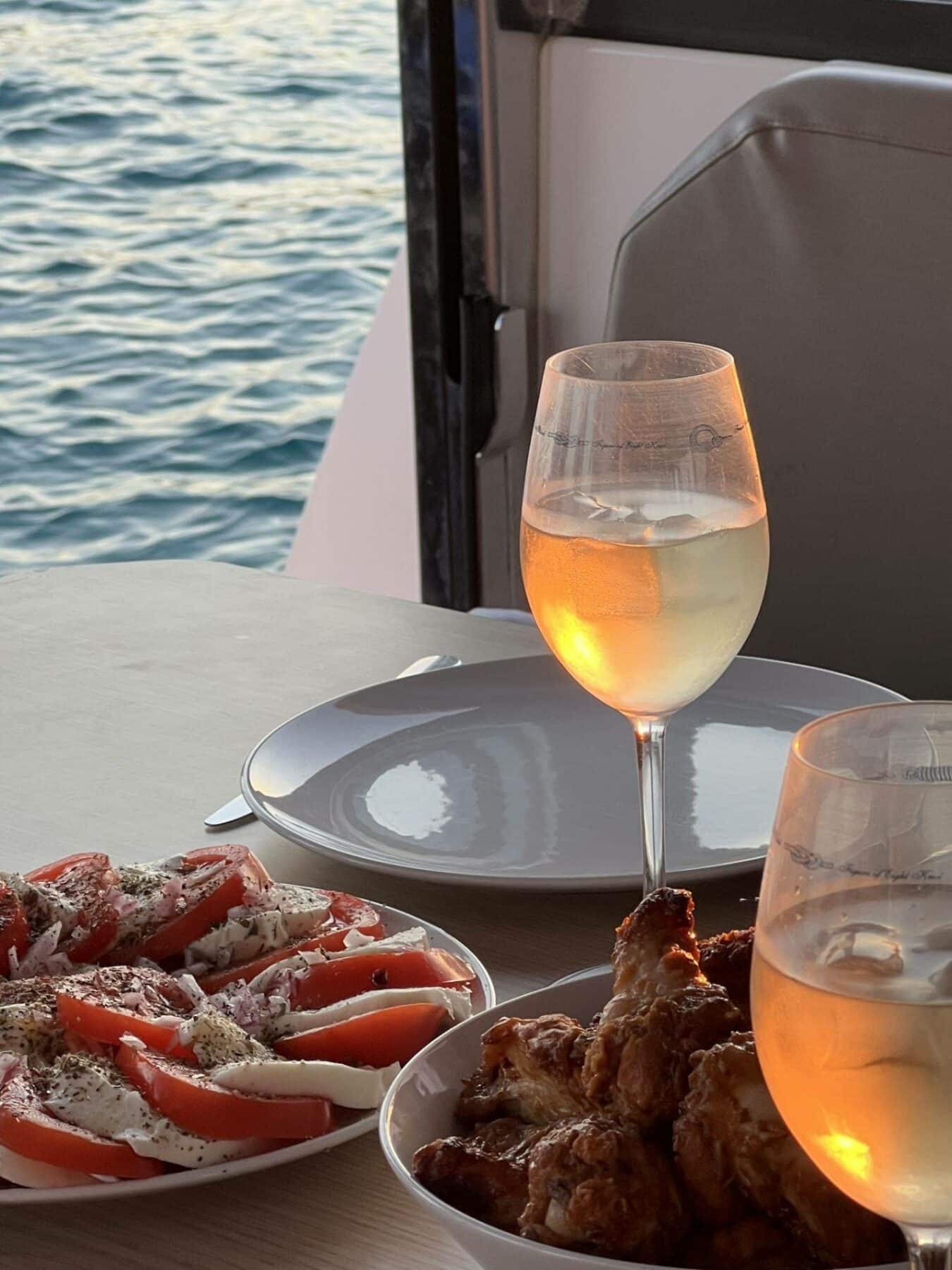 Eine entspannende Mahlzeit am Meer mit einem Glas Roséwein, Tellern mit Tomaten-Mozzarella-Salat und gebratenem Hähnchen vor der ruhigen Meereskulisse während eines Segeltörns bei Sonnenuntergang.