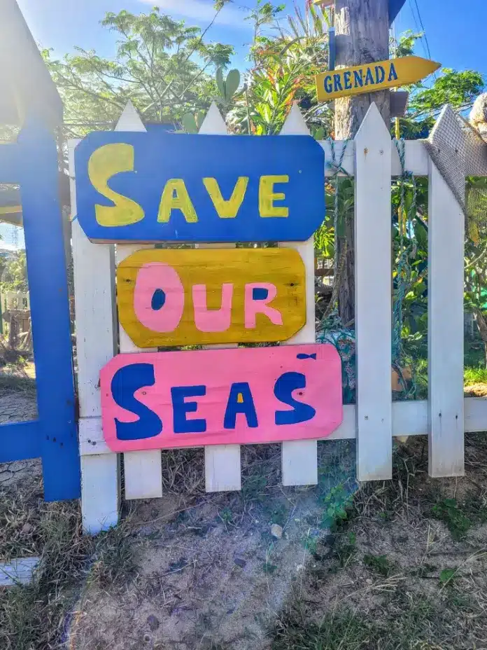 Buntes "Rettet unsere Meere"-Schild an einem weißen Lattenzaun mit einem Richtungspfeil mit der Aufschrift "Grenada", der von einer Segelyacht nach rechts zeigt, umgeben von üppigem Grün