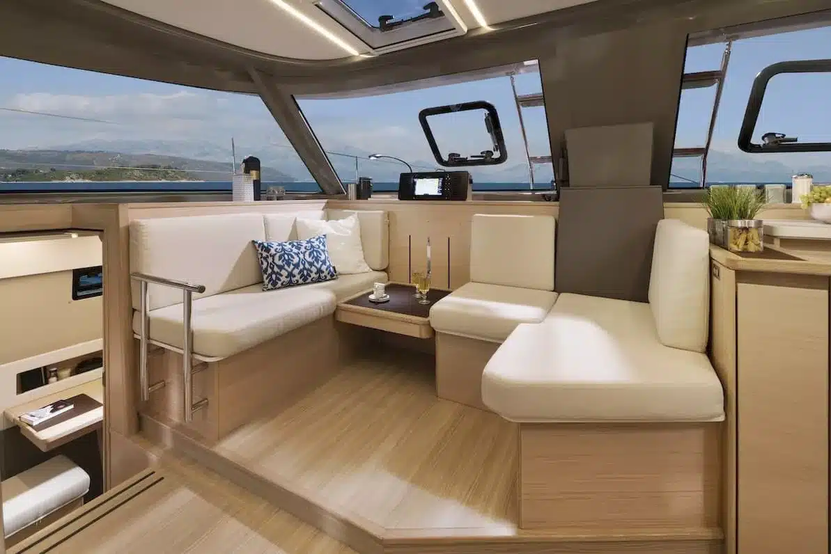 Luxuriöses Yacht-Interieur mit beigen Ledersitzen, Holzboden und großen Fenstern mit Blick auf die Küste. Dekorative Kissen und Getränke schmücken den Raum, perfekt für ein Segeltörn-Abenteuer.