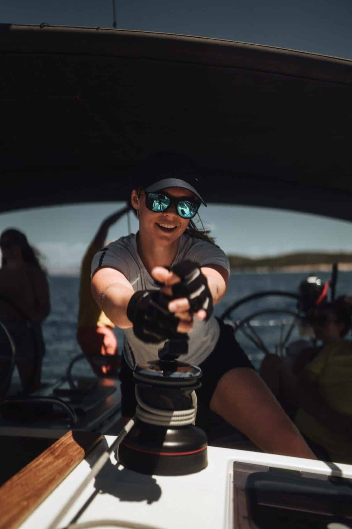 Eine Frau mit Mütze und Sonnenbrille bedient die Winde eines Segelboots. Ihr Spiegelbild ist in der Sonnenbrille zu sehen, die während ihres Segelurlaubs vom hellen Sonnenlicht angestrahlt wird.