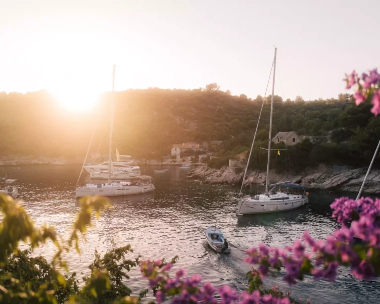 Blick auf den Sonnenuntergang über einer ruhigen Bucht mit einer Segelyacht, die in der Nähe einer felsigen Küste vor Anker liegt, im Vordergrund eingerahmt von rosa Blumen.