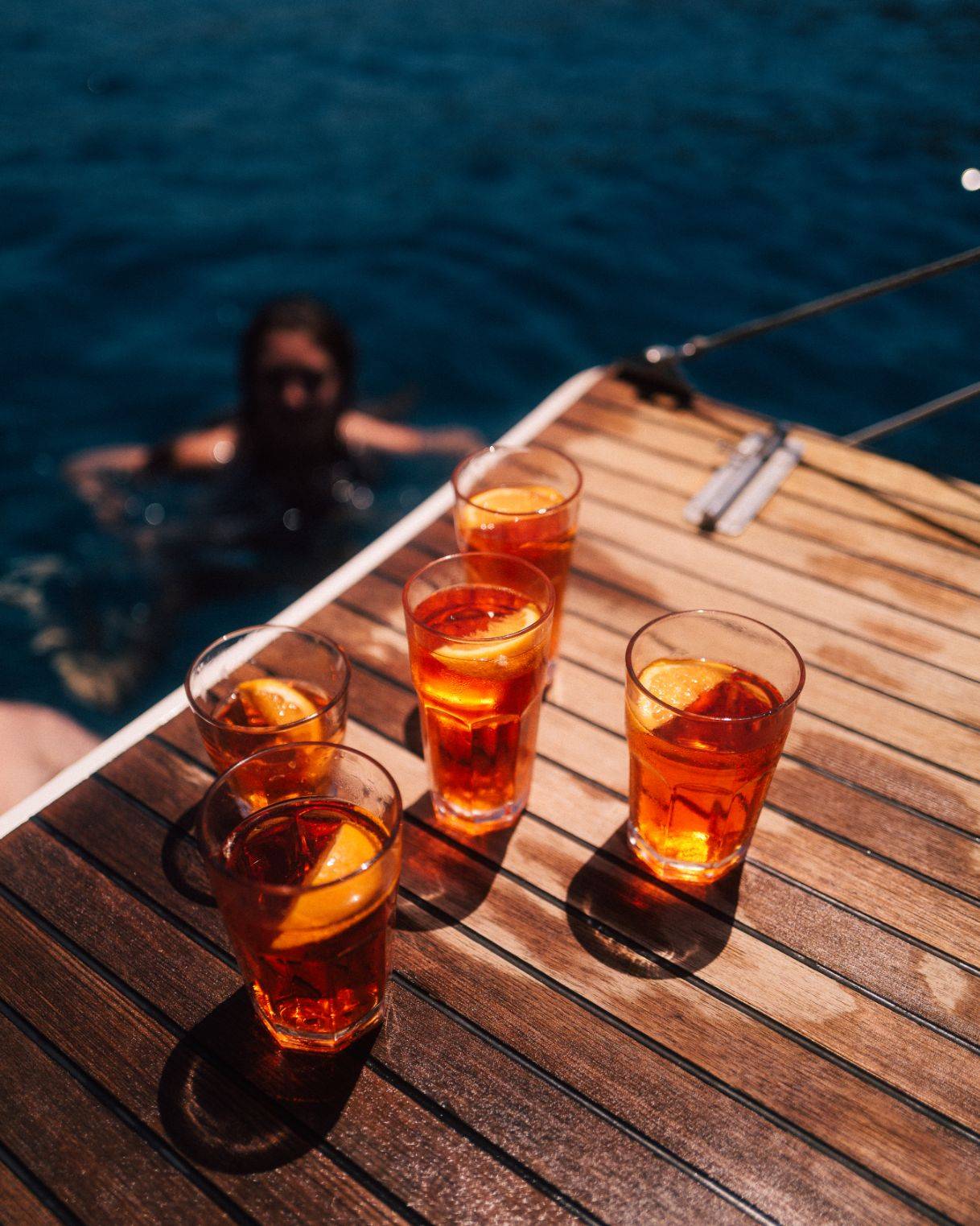 Vier Gläser Eistee mit Zitronenscheiben auf dem Holzdeck einer Segelyacht, mit Blick auf das klare blaue Wasser und einer schwimmenden Person im Hintergrund.