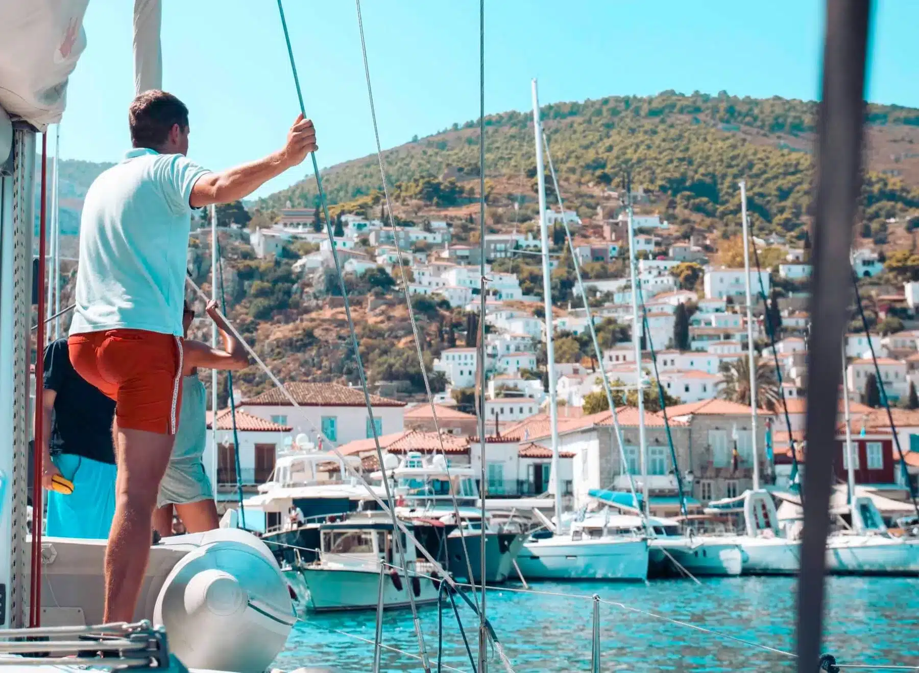 Ein Mann auf einem Segelboot hält ein Seil und blickt auf eine malerische Küstenstadt mit weißen Gebäuden und blauem Wasser. Andere Boote sind in der Nähe festgemacht, während er seinen Segelurlaub verbringt.