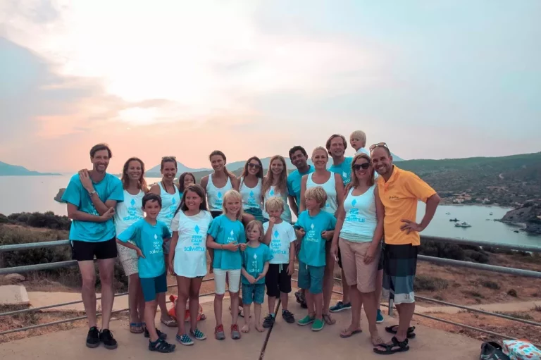 Eine große Familiengruppe in Freizeitkleidung und passenden blauen Hemden steht zusammen auf einer Segelyacht mit einer malerischen Sonnenuntergangslandschaft und ruhigem Meer im Hintergrund.