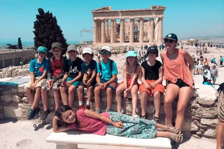 Eine Gruppe von Kindern sitzt auf einer Steinbank vor dem Parthenon in Athen, Griechenland. Ein Kind liegt davor und lächelt in die Kamera, an einem sonnigen Tag mit klarem blauen Himmel.