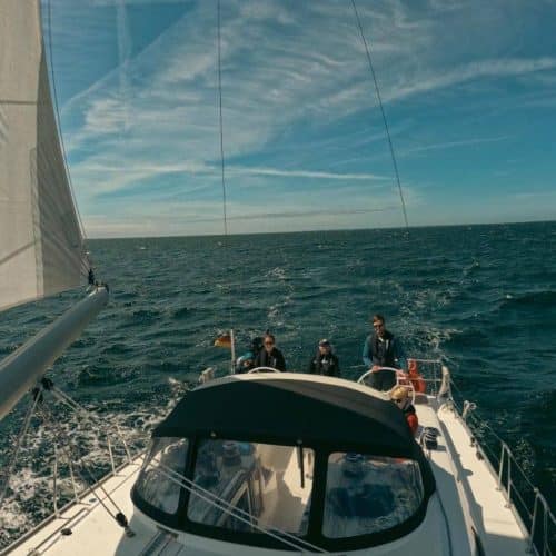 Eine Gruppe von Menschen genießt einen Segelurlaub an einem sonnigen Tag, mit klarem Himmel und unruhigem blauen Wasser rundherum, gesehen vom Heck der Segelyacht.