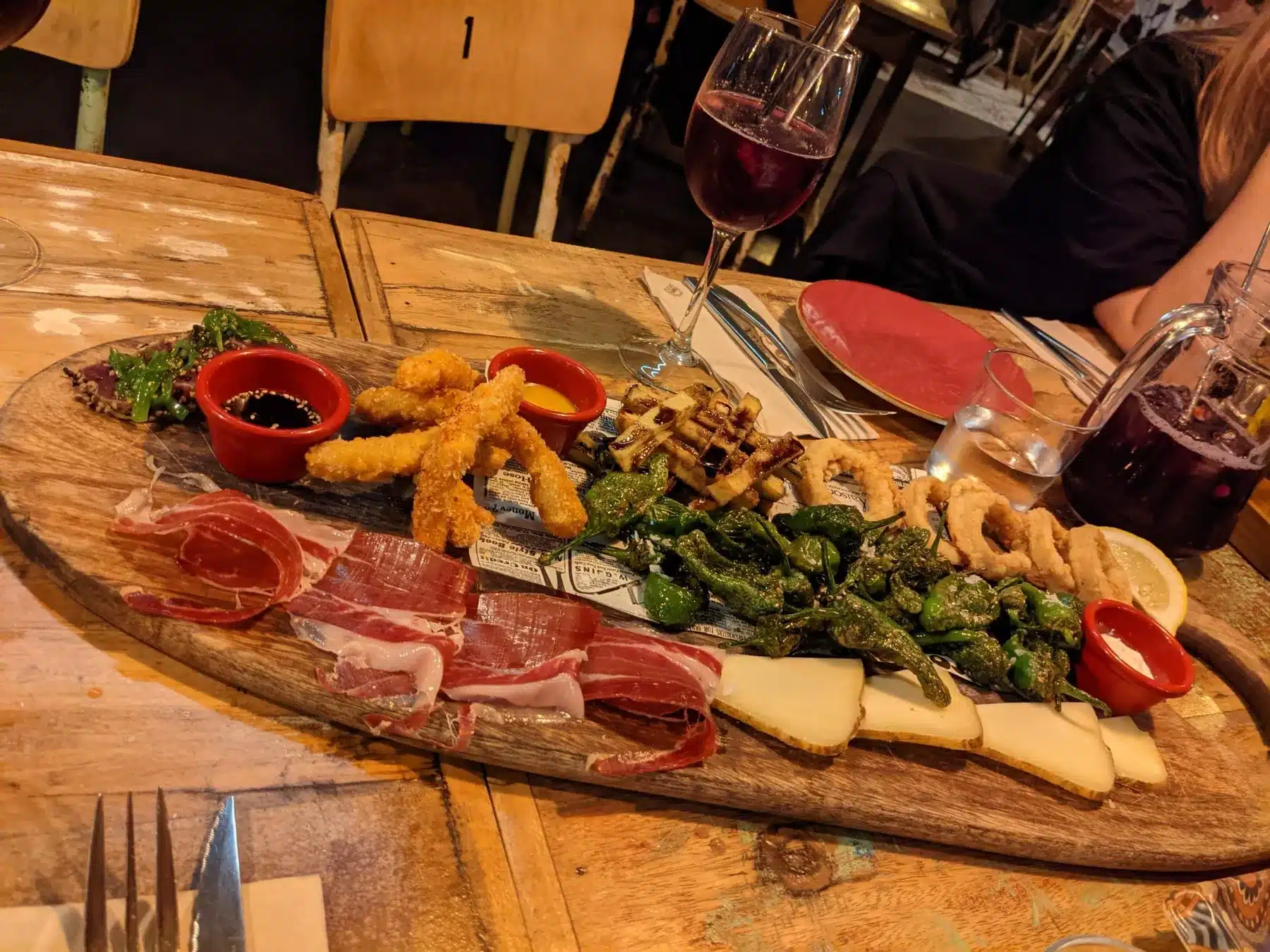 Eine Holzplatte gefüllt mit verschiedenen Tapas, darunter Wurstwaren, Käse, frittierte Meeresfrüchte und Gemüse, serviert mit Gläsern Rotwein auf einem Restauranttisch während eines Segeltörn.