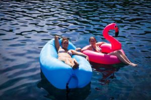 Zwei Menschen entspannen auf aufblasbaren Schwimmkörpern, einer in Form eines blauen Donuts und der andere in Form eines rosa Flamingos, während sie auf einer sonnenbeschienenen, klaren, blauen Wasserfläche segeln.