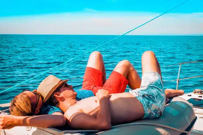 Ein Paar entspannt sich auf einem Segelboot. Sie liegen Rücken an Rücken mit hochgelegten Beinen, tragen Sonnenbrillen und Hüte und im Hintergrund das blaue Meer und der Himmel.