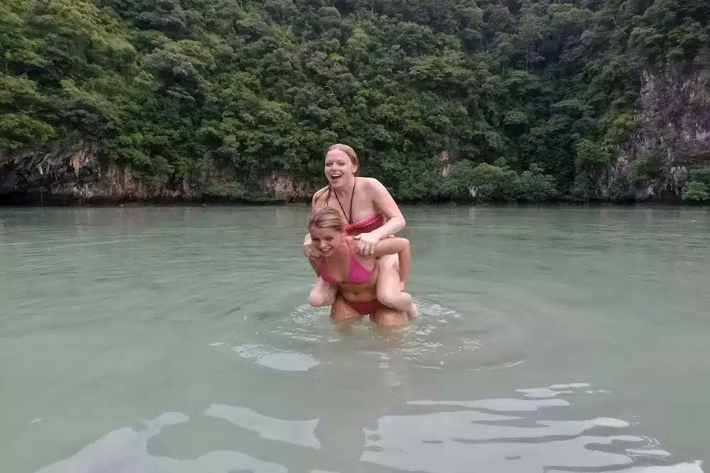 Zwei Frauen lachen fröhlich, während eine die andere Huckepack durch einen ruhigen Fluss mit üppigen grünen Hügeln und einer Segelyacht im Hintergrund trägt.