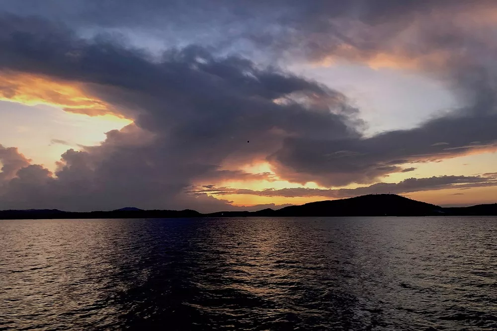 Ein dramatischer Sonnenuntergang über einem ruhigen See mit Hügelsilhouetten am Horizont und Wolken, die in warmen Gold- und Orangetönen vor einem dunkelblauen Himmel während eines Segelurlaubs erstrahlen.