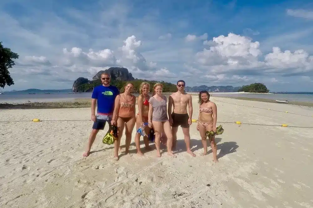 Eine Gruppe von sieben Erwachsenen steht lächelnd an einem sonnigen Strand mit klarem Himmel. Der Strand hat hellen Sand und im Hintergrund ist eine malerische Insel zu sehen. Sie tragen Sommerkleidung und halten Schnorchelausrüstung in der Hand