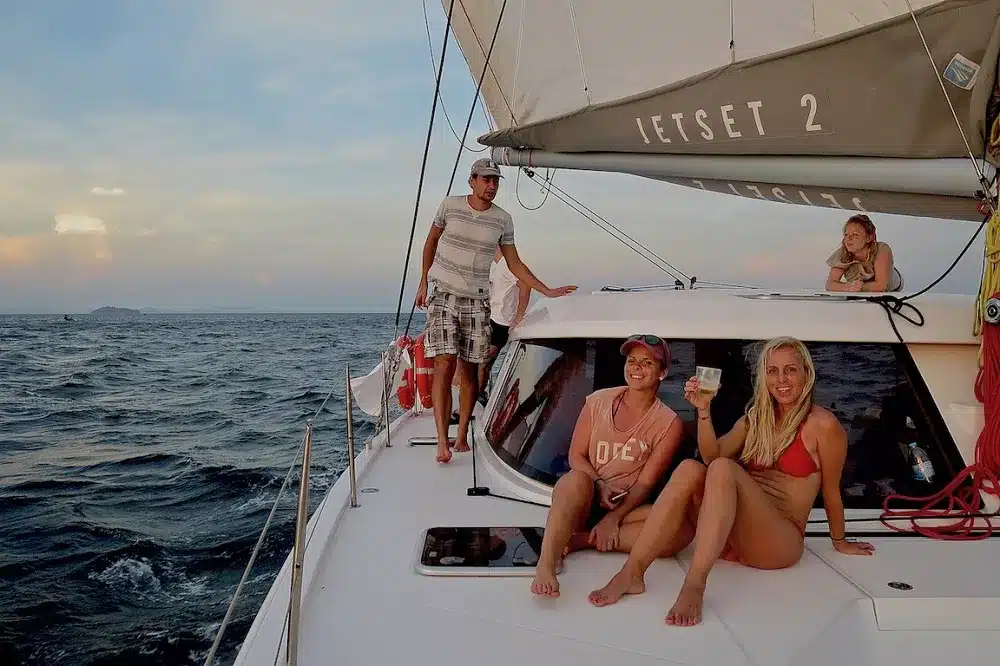 Drei Menschen genießen eine Segelreise auf einer Yacht namens "Jetset 2", mit zwei Frauen sitzen und lächeln an der Vorderseite des Bootes, und ein Mann steht am Mast, Ozean