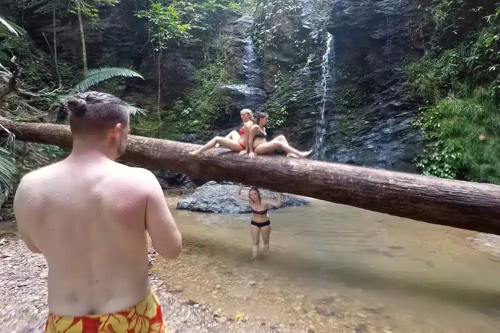 Eine Gruppe von drei Personen genießt einen natürlichen Wasserfall. Ein Mann sitzt auf einem umgestürzten Baum, ein anderer steht im Wasser und macht ein Foto, und eine Frau posiert verspielt auf dem Baumstamm. Saftiges Grün