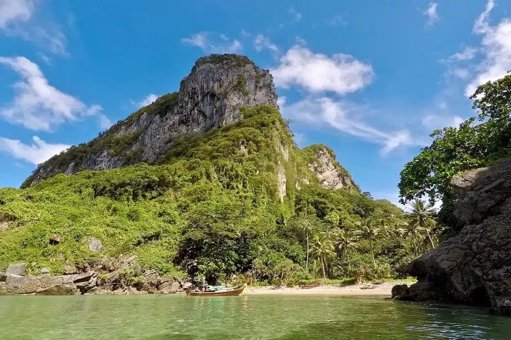 Ein ruhiger tropischer Strand mit einer hoch aufragenden Kalksteinklippe, die von üppigem Grün unter einem klaren blauen Himmel bedeckt ist, und einer kleinen Segelyacht, die in Ufernähe vor Anker liegt.