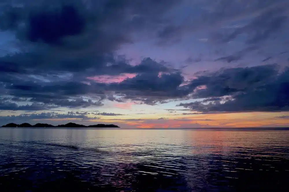 Ein heiterer Sonnenuntergang über einem ruhigen Meer mit vereinzelten Wolken, der einen Farbverlauf von tiefem Blau zu leuchtendem Orange zeigt, und schattigen Inseln in der Ferne während eines friedlichen Segelurlaubs.
