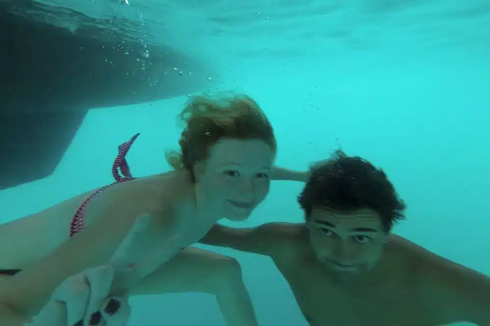 Zwei Menschen schwimmen unter Wasser, einer lächelt in die Kamera und macht ein Peace-Zeichen, während der andere zusieht. Ihre Ausdrücke sind verspielt und sie scheinen das klare Wasser um sie herum zu genießen während ihrer