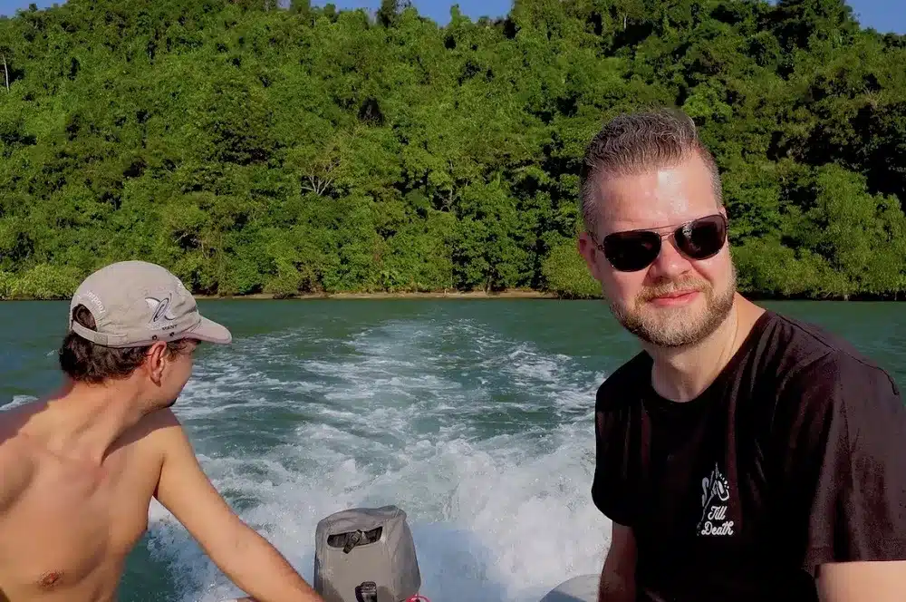 Zwei Männer auf einem Segelboot, einer steuert mit dem Außenbordmotor, der andere lächelt in die Kamera, im Hintergrund ein üppiger grüner Wald und plätscherndes Wasser.
