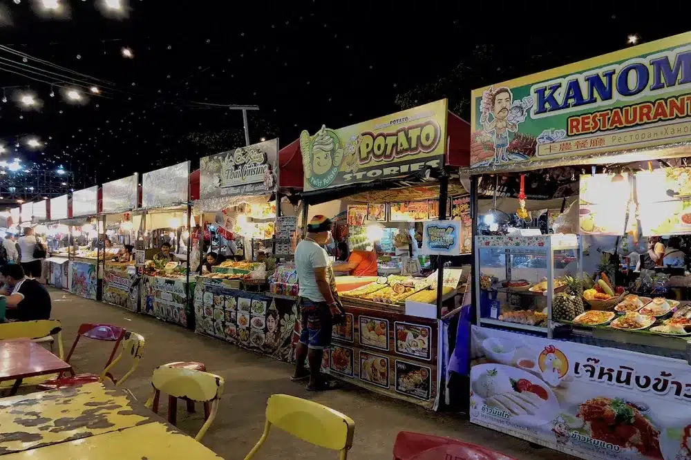 Eine geschäftige Nachtmarktszene mit verschiedenen Essensständen, darunter „Potato Stories“ und „Kanom Restaurant“, die mit farbenfrohen Schildern ausgestattet sind und unter einem dunklen, von Lichterketten erhellten Himmel abwechslungsreiche Speisen anbieten.
