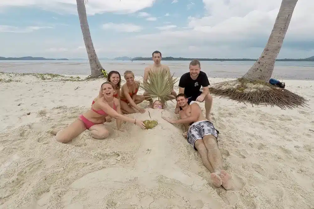 Eine Gruppe von sieben Personen, die nach einem Segelabenteuer Spaß an einem Sandstrand haben; einer ist spielerisch bis zum Hals im Sand vergraben und trägt einen Hut aus Palmblättern, umgeben von den anderen, die lächeln und