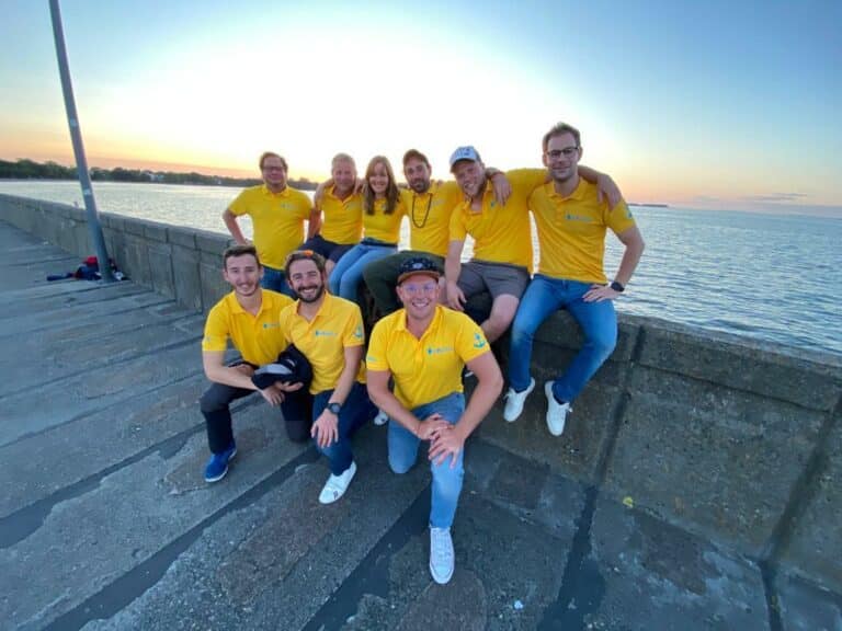Eine Gruppe von acht Personen in passenden gelben Hemden posiert fröhlich bei Sonnenuntergang auf einer Segelyacht.