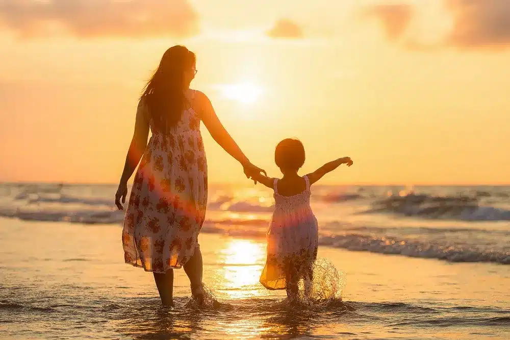 Eine Mutter und ihre Tochter gehen auf ihrer Segelreise Händchen haltend bei Sonnenuntergang durch das seichte Meerwasser und heben sich als Silhouette vor dem goldenen Himmel ab.