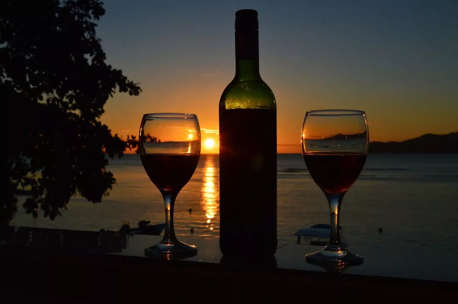 Zwei Gläser Wein und eine Flasche auf einem Geländer, die sich vor dem Hintergrund eines leuchtenden Sonnenuntergangs über einem ruhigen See abzeichnen, in der Ferne sind Segelboote zu sehen.