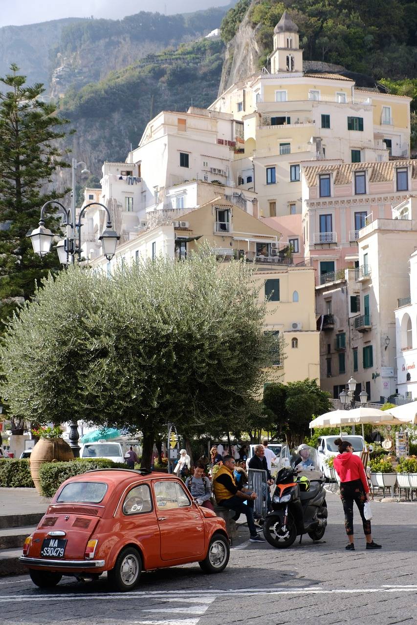 Eine belebte Straßenszene in einer italienischen Stadt mit einem roten Oldtimer im Vordergrund, Menschen auf Motorrollern und Fußgängern vor der Kulisse weißer Gebäude und eines felsigen Hügels.