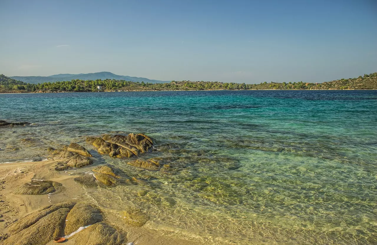 Eine ruhige Strandlandschaft mit kristallklarem, türkisfarbenem Wasser und felsigen Küstenlinien, vor der Kulisse üppig grüner Hügel unter einem klaren, blauen Himmel, perfekt für einen Segeltörn.