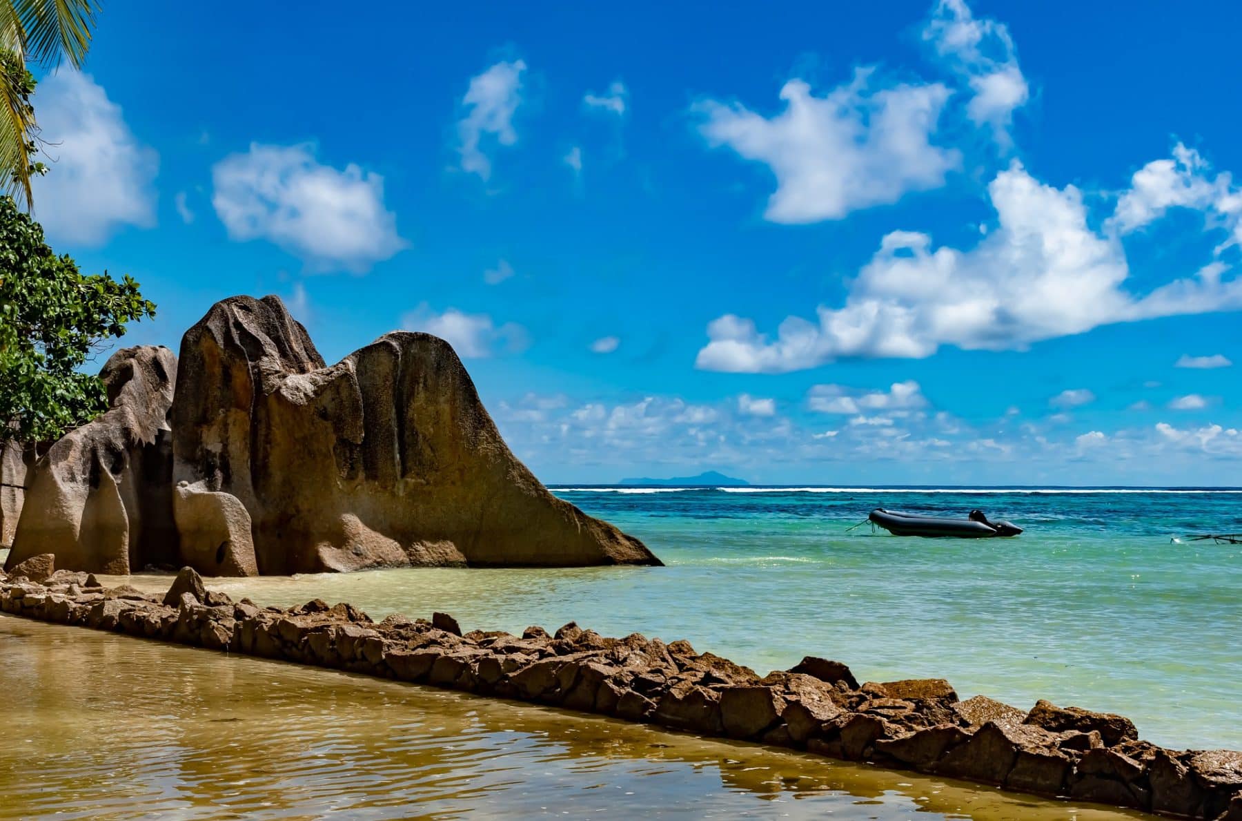 Ein malerischer tropischer Strand mit großen Granitfelsen am Ufer, klarem türkisfarbenem Wasser und einem blauen Himmel mit flauschigen Wolken. Eine Segelyacht schwimmt nahe dem Horizont.