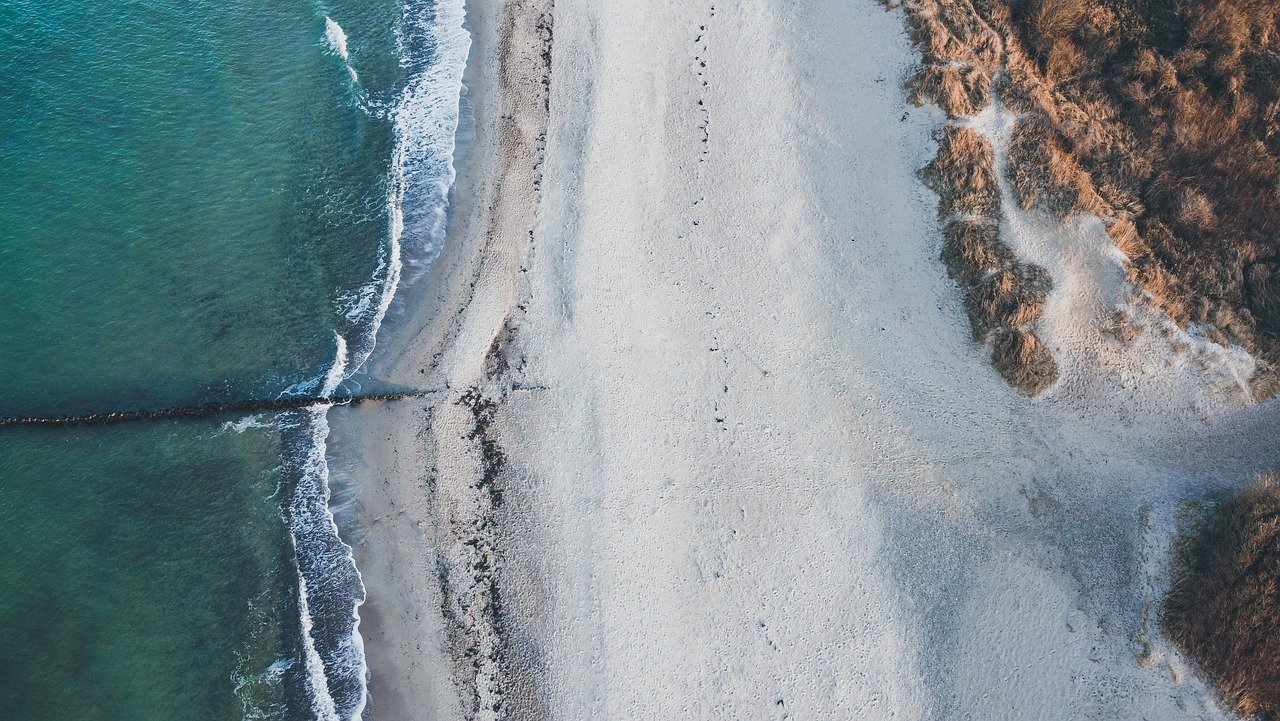 Luftaufnahme einer Küste, an der ein Sandstrand auf das blaue Meer trifft, getrennt durch die sanften Wellen eines Segeltörns, was von oben eine ruhige, natürliche Grenze veranschaulicht.