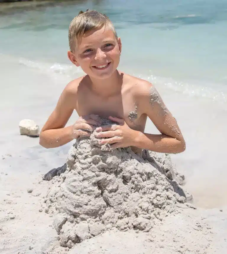 Ein kleiner Junge mit einem fröhlichen Lächeln, bedeckt mit Sand, kniet an einem Strand und baut eine Sandburg. Hinter ihm ist das klare blaue Wasser voller Menschen zu sehen, die ihren Segelurlaub genießen.