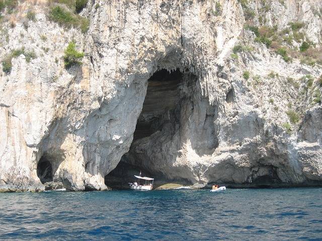 Ein Segelboot und ein Kajak fahren an einem sonnigen Tag mit klarem Himmel in eine große Höhle in einer zerklüfteten Felswand über tiefblauem Wasser.